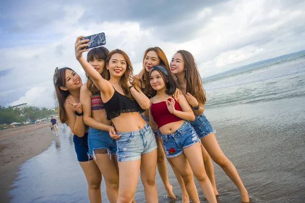lifestyle beach portrait of Asian Korean and Chinese women, group of happy beautiful young girlfriends taking selfie picture together with mobile phone smiling cheerful enjoying girls holidays trip