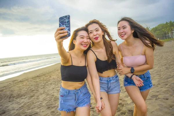 lifestyle beach portrait of Asian Korean and Chinese women, group of happy beautiful young girlfriends taking selfie picture together with mobile phone smiling cheerful enjoying girls holidays trip