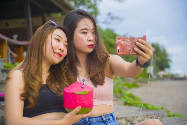 lifestyle portrait of two young happy and attractive Asian Chinese and Korean women hanging out , girlfriends enjoying holidays trip in tropical resort taking selfie picture with mobile phone