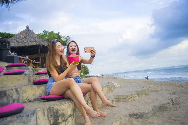 lifestyle portrait of two young happy and attractive Asian Chinese and Korean women hanging out , girlfriends enjoying holidays trip in tropical resort taking selfie picture with mobile phone