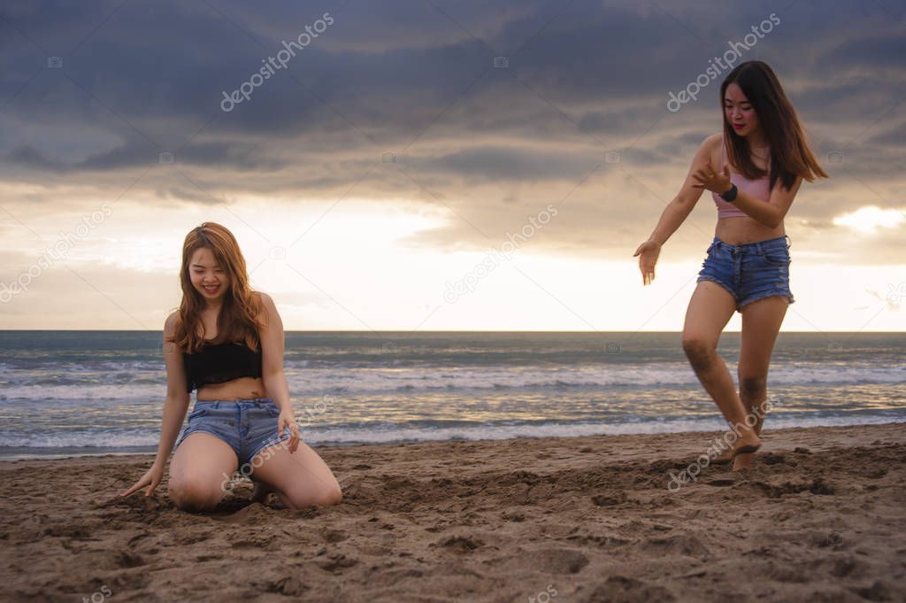 two happy and attractive young Asian Chinese women girlfriends or sisters having fun playing with sand on sunset beach in beautiful light enjoying summer holidays travel together carefree