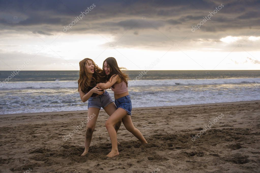 two happy and attractive young Asian Chinese women girlfriends or sisters having fun playing wrestling on sunset beach in beautiful light enjoying summer holidays travel together carefree