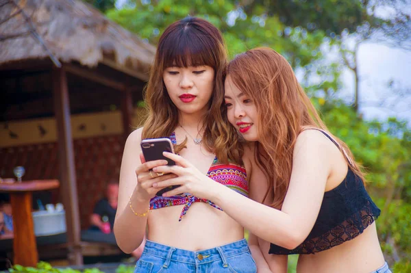 two happy and pretty Asian Korean girlfriends enjoying Summer holidays trip together at tropical beach using internet on mobile phone relaxed and cheerful in women friendship concept