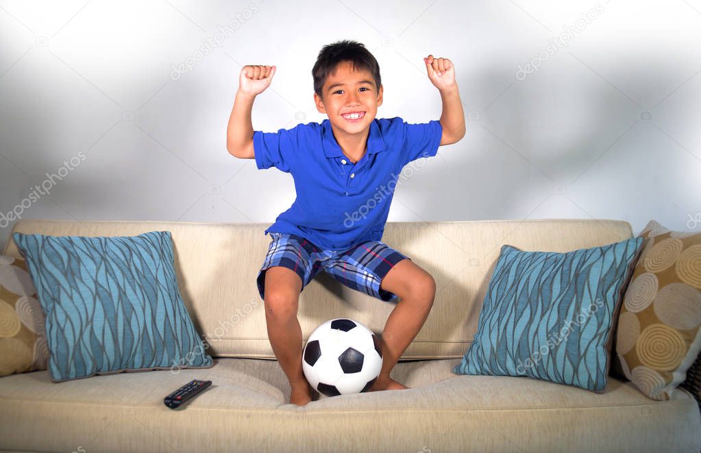 young hispanic boy happy and excited watching football game on television jumping excited on home living couch , the kid celebrating scoring goal gesturing cheerful as soccer crazy fan