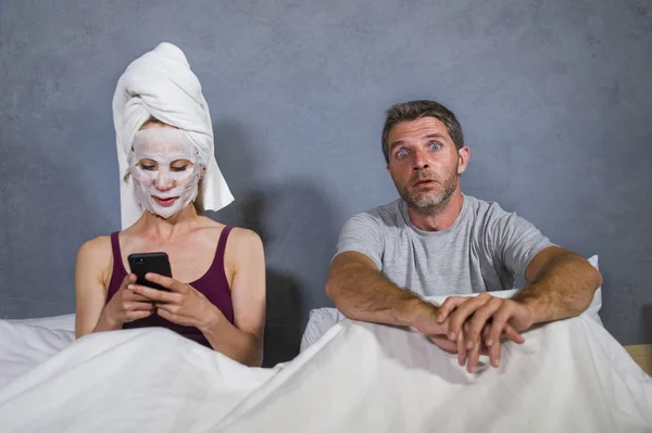 funny lifestyle portrait of eccentric housewife with makeup facial mask and towel using mobile phone in bed and husband in desperate face expression in weird man woman relationship concept