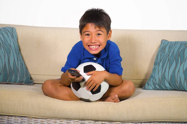 lifestyle portrait at home of young 7 or 8 years old boy holding soccer ball watching excited and nervous football game on television sitting at living sofa couch suffering and enjoying sport emotion