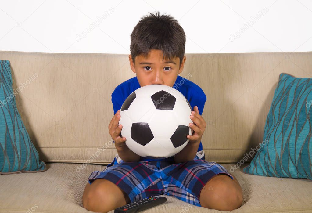lifestyle portrait at home of young 7 or 8 years old boy holding soccer ball watching excited and nervous football game on television sitting at living sofa couch suffering and enjoying sport emotion