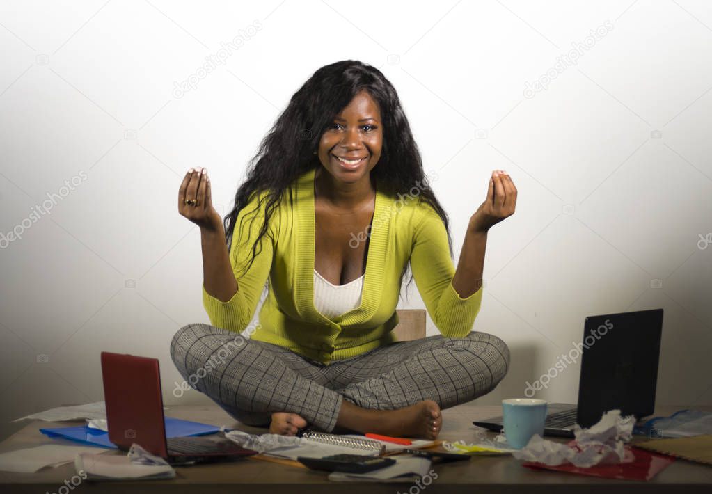 young happy and attractive African American business woman doing yoga sitting at office messy desk full of paperwork smiling relaxed and carefree in anti-stress therapy concept isolated on white 