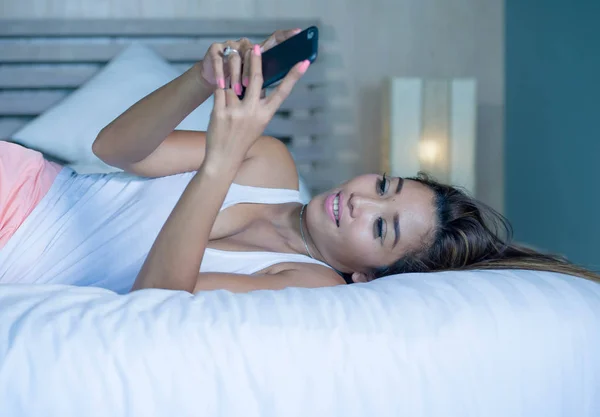 young attractive and happy Asian woman lying relaxed on bed using internet social media app in mobile phone smiling sending text online dating in communication lifestyle concept