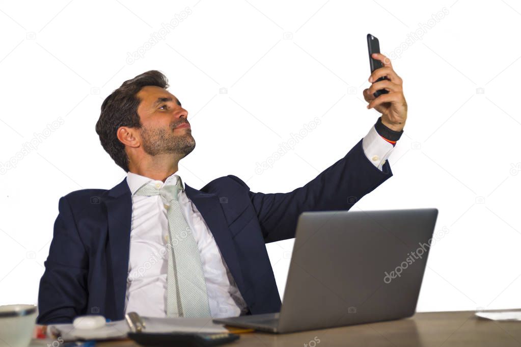 young attractive and confident businessman in suit working at corporate company office computer desk taking selfie portrait picture with mobile phone camera feeling cool and successful