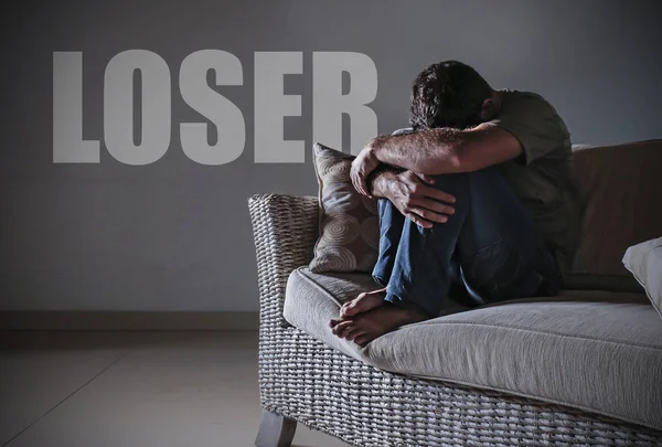 young desperate sad and frustrated man grieving at home sofa couch suffering depression problem and anxiety crisis crying with the word loser on the wall feeling miserable and lost