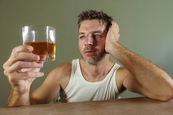 young wasted and depressed alcohol addict man in dirty singlet drinking glass of whiskey feeling desperate suffering alcoholism problem and booze addiction on isolated background in alcoholic concept