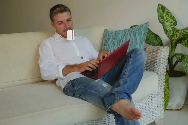 young handsome and attractive happy man doing internet shopping online with credit card sitting relaxed at home sofa couch smiling cheerful in electronic business and commerce concept