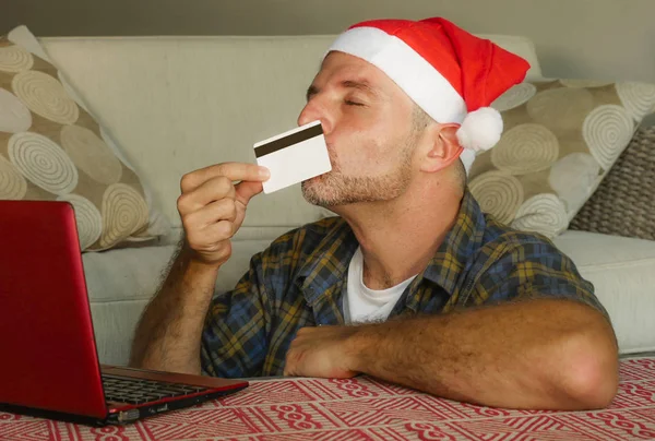サンタの帽子を自宅で若いハンサムで魅力的な幸せな男は クレジット カードを購入し クリスマス プレゼントを コマースの概念を支払うことを使用してラップトップ コンピューターとインターネットにオンライン ショッピング — ストック写真