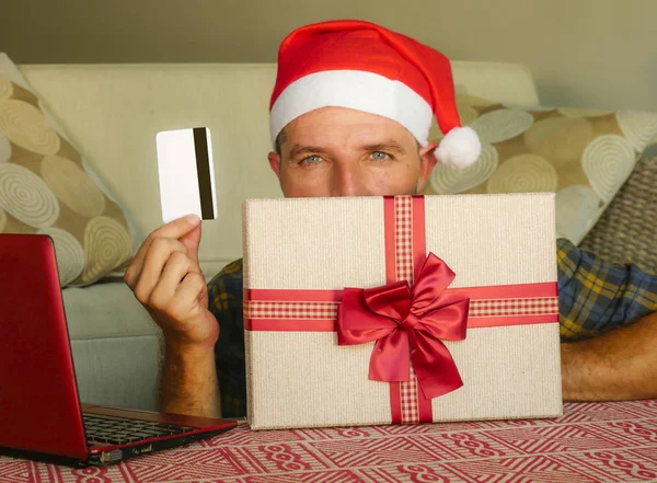 年轻英俊和有吸引力的幸福的人在圣诞老人的帽子在家里沙发使用笔记本电脑和信用卡持有圣诞礼品盒网上购物电子商务和互联网业务概念 — 图库照片
