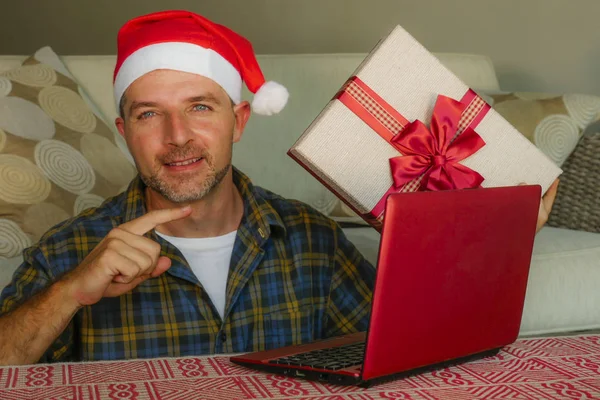 young handsome and attractive happy man in Santa hat at home online shopping on internet with laptop computer holding present box buying Christmas present in ecommerce concept