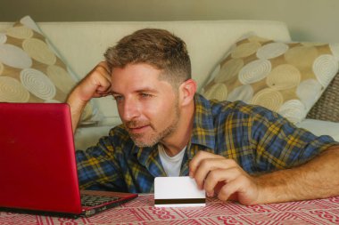 kredi kartı kullanarak dizüstü bilgisayar online alışveriş internet iş ve e-ticaret kavramı içinde neşeli gülümseyen koltuk kanepe evde oturan tutan genç yakışıklı ve mutlu adam