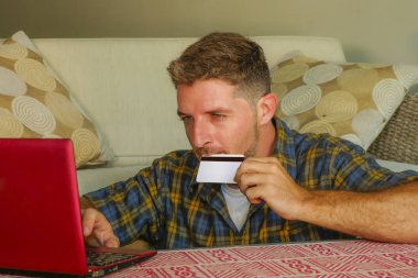 kredi kartı kullanarak dizüstü bilgisayar online alışveriş internet iş ve e-ticaret kavramı içinde neşeli gülümseyen koltuk kanepe evde oturan tutan genç yakışıklı ve mutlu adam