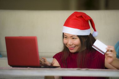 Genç güzel ve mutlu Asyalı Çinli kadın Noel Noel Baba şapkalı kredi kartı alışveriş online gülümseyen Internet için dizüstü bilgisayar kullanan evde kanepe oturma odası heyecanlı holding