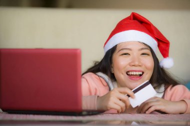 Santa şapka holding kredi kartı ve Noel giyen genç güzel ve mutlu Asyalı Koreli kadın online neşeli gülümseyen evde alışveriş dizüstü bilgisayar için Internet kullanan kutusu sunmak