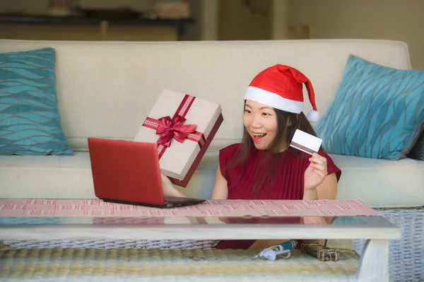 年轻美丽和快乐的亚洲韩国妇女在圣诞老人帽子持有信用卡和圣诞礼物盒使用笔记本电脑上网购物微笑购买礼物从家里 — 图库照片