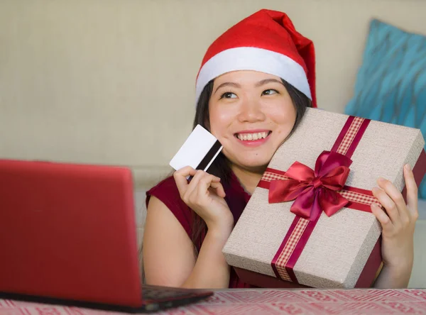 年轻美丽和快乐的亚洲韩国妇女在圣诞老人帽子持有信用卡和圣诞礼物盒使用笔记本电脑上网购物微笑购买礼物从家里 — 图库照片
