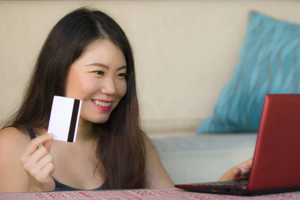 lifestyle portrait of young beautiful and happy Asian Chinese woman holding credit card banking and online shopping with laptop computer at home couch in internet business and e.commerce concept