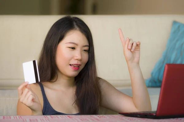 lifestyle portrait of young beautiful and happy Asian Korean woman holding credit card banking and online shopping with laptop computer at home couch in internet business and e.commerce concept