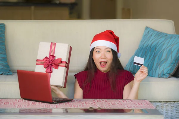 年轻美丽和快乐的亚洲女性在圣诞老人帽子拿着信用卡和圣诞节礼物盒使用笔记本电脑上网购物微笑购买礼物从家里 — 图库照片