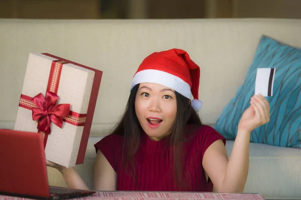 年轻美丽快乐的亚洲中国妇女拿着圣诞老人的帽子和圣诞礼物盒使用笔记本电脑上网购物微笑愉快在家里 — 图库照片