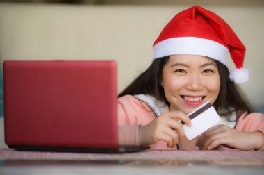 Santa şapka holding kredi kartı ve Noel giyen genç güzel ve mutlu Asyalı Çinli kadın online neşeli gülümseyen evde alışveriş dizüstü bilgisayar için Internet kullanan kutusu sunmak