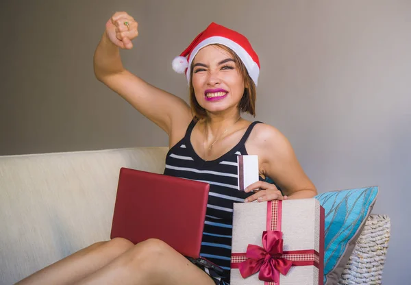 young happy and attractive woman relaxed at apartment couch in Christmas hat using laptop computer buying present with credit card smiling cheerful in online shopping and internet commerce