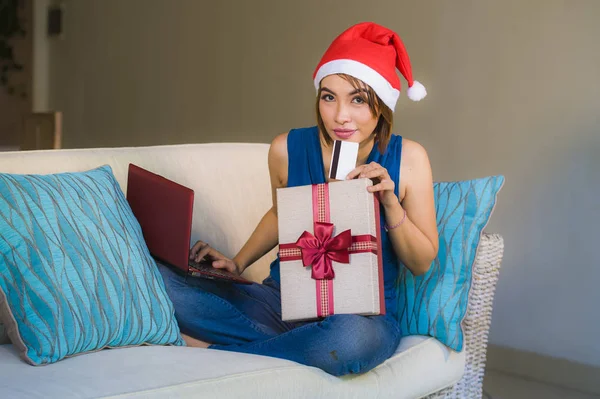 年轻的快乐和美丽的女孩放松在家里的沙发上圣诞老人帽子使用笔记本电脑支付圣诞礼物与信用卡微笑在网上购物和互联网商务 — 图库照片