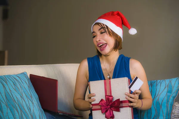 年轻的快乐和美丽的女孩放松在家里的沙发上圣诞老人帽子使用笔记本电脑支付圣诞礼物与信用卡微笑在网上购物和互联网商务 — 图库照片