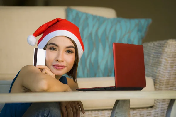 young happy and beautiful girl relaxed at home couch in Santa hat using laptop computer paying for Christmas present with credit card smiling in online shopping and internet commerce