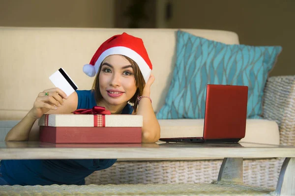 年轻快乐和美丽的女人放松在家里的沙发上圣诞老人帽子使用笔记本电脑支付圣诞礼物与信用卡微笑在网上购物和互联网商务 — 图库照片