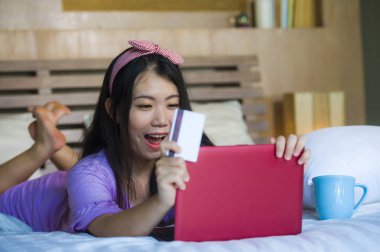 kredi kartı internet bankacılığı kullanan genç güzel ve mutlu Asya Japon kadın evde yatakta neşeli alışveriş online e-ticaret ve yaşam tarzı kavramında satın alma gülümseyen dizüstü bilgisayar üzerinde