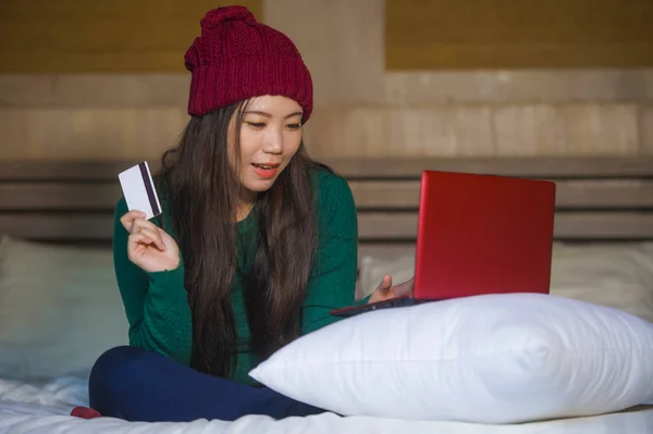 生活方式家庭的年轻美丽和快乐的亚洲女孩在冬季帽子坐轻松地在床上拿着信用卡使用笔记本电脑网上银行和网上购物 — 图库照片