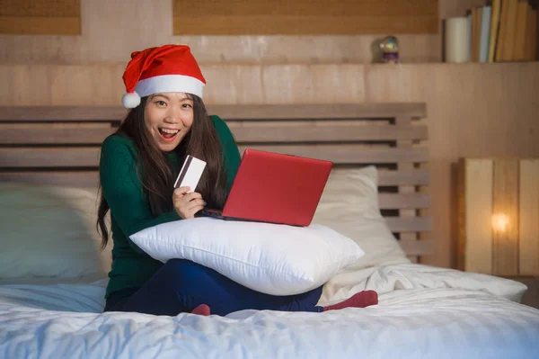 年轻快乐和美丽的亚洲日本妇女在圣诞老人的圣诞帽使用信用卡和笔记本电脑在线购物圣诞礼物和礼物坐在快乐和兴奋的床上 — 图库照片