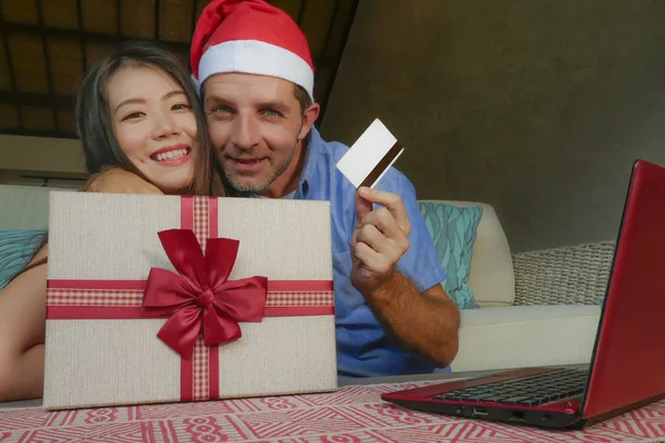 young happy and excited mixed ethnicity couple with Asian Chinese woman and Caucasian man in Christmas Santa hat online shopping x-mas presents with credit card and laptop computer at home