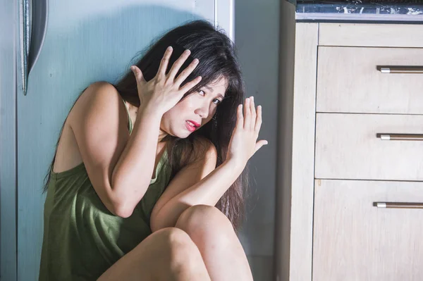 young scared and helpless Asian Korean woman horrified suffering domestic violence victim of abuse from drunk alcoholic husband at home kitchen floor crying desperate in fear and panic