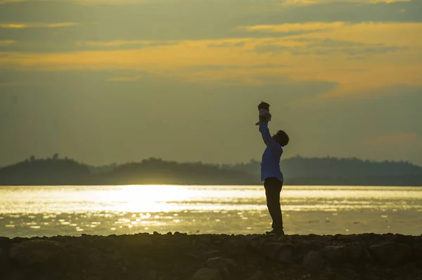 Silhouette of young happy man with his little son or daughter in his arms , the father raising up the baby in front of the sea at beach boardwalk enjoying sunset during holidays trip
