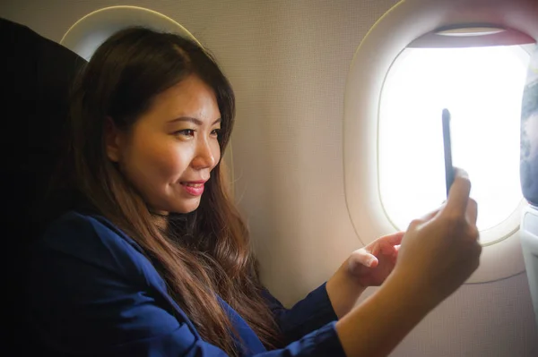 young happy and beautiful Asian Chinese woman traveling for business inside airplane cabin smiling cheerful using mobile phone taking selfie self portrait picture in air travel and tourism concept