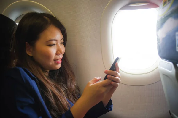 lifestyle portrait of young happy and beautiful Asian Chinese woman traveling for business inside airplane cabin smiling cheerful using mobile phone in wifi and internet on flight concept