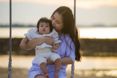 yaşam tarzı açık havada birlikte yaz plaj salıncakta, anne ve küçük kızı sevgi kavramı günbatımı sallanan kız bebek tutan genç tatlı ve mutlu Asyalı Çinli kadın portresi 