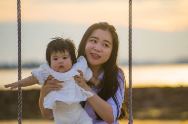 yaşam tarzı açık havada birlikte yaz plaj salıncakta, anne ve küçük kızı sevgi kavramı günbatımı sallanan kız bebek tutan genç tatlı ve mutlu Asyalı Çinli kadın portresi 