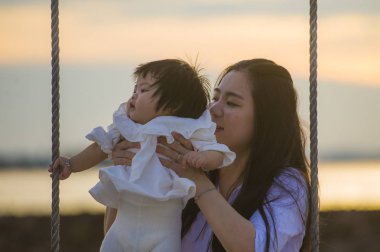 yaşam tarzı açık havada birlikte yaz plaj salıncakta, anne ve küçük kızı sevgi kavramı günbatımı sallanan kız bebek tutan genç tatlı ve mutlu Asyalı Çinli kadın portresi 