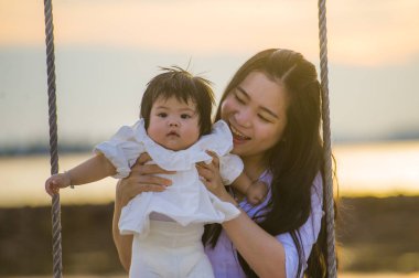 yaşam tarzı açık havada birlikte yaz plaj salıncakta, anne ve küçük kızı sevgi kavramı günbatımı sallanan kız bebek tutan genç tatlı ve mutlu Asyalı Çinli kadın portresi 