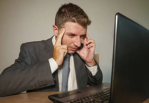 young frustrated and stressed businessman in suit and tie working overwhelmed at office laptop computer desk suffering headache and migraine feeling sick in corporate job problem