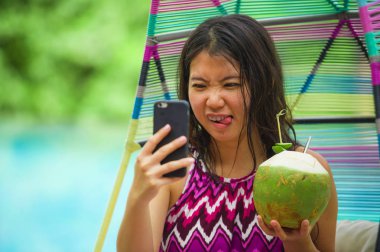 Genç güzel ve mutlu Asyalı Koreli turist kadın içme Hindistan cevizi suyu selfie fotoğrafını cep telefonu ile lüks tropik ada resort yüzme havuzunun keyfini yaz tatili 